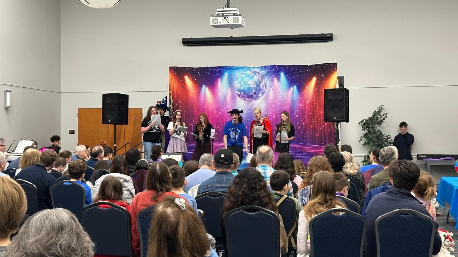 Purim Shpiel performance on stage with colorful disco backdrop and packed audience