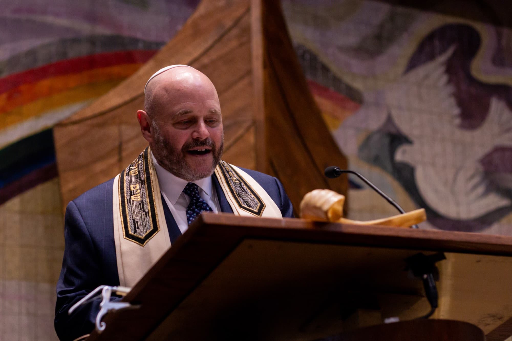 Rabbi Doug speaking passionately from the podium with the colorful sanctuary mural behind him