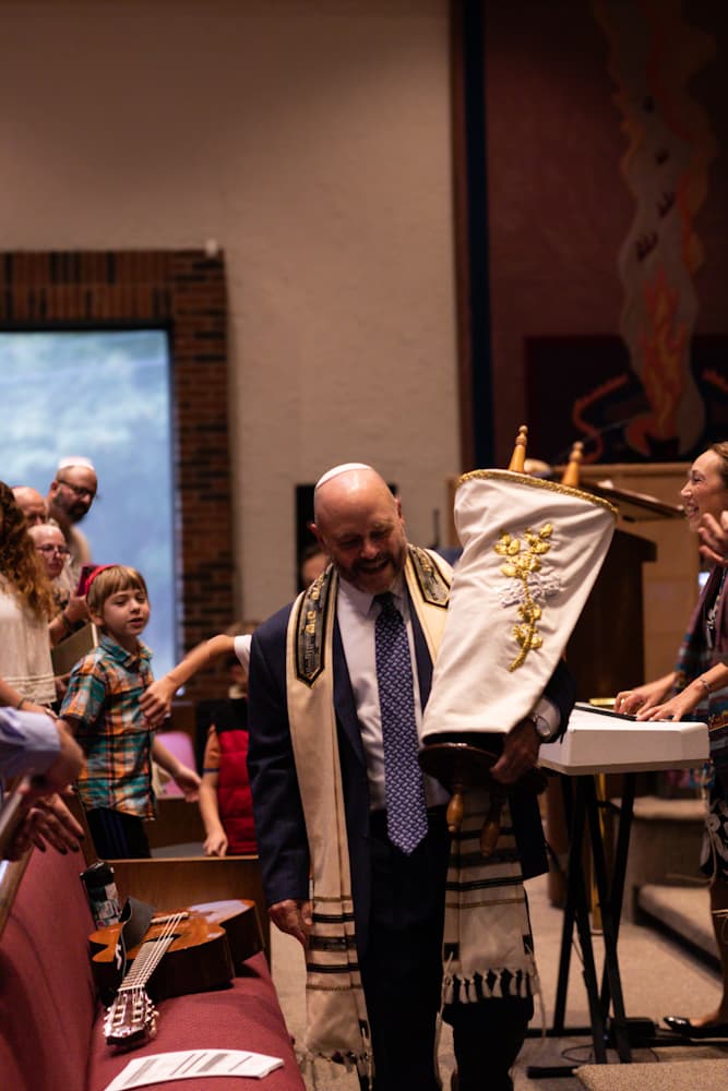 Rabbi Doug holding the Torah scroll with a child and community members in the sanctuary with colorful mural backdrop
